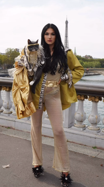 Bagel carried on the shoulder of a model in gold couture on a Paris bridge, with the Eiffel Tower behind them